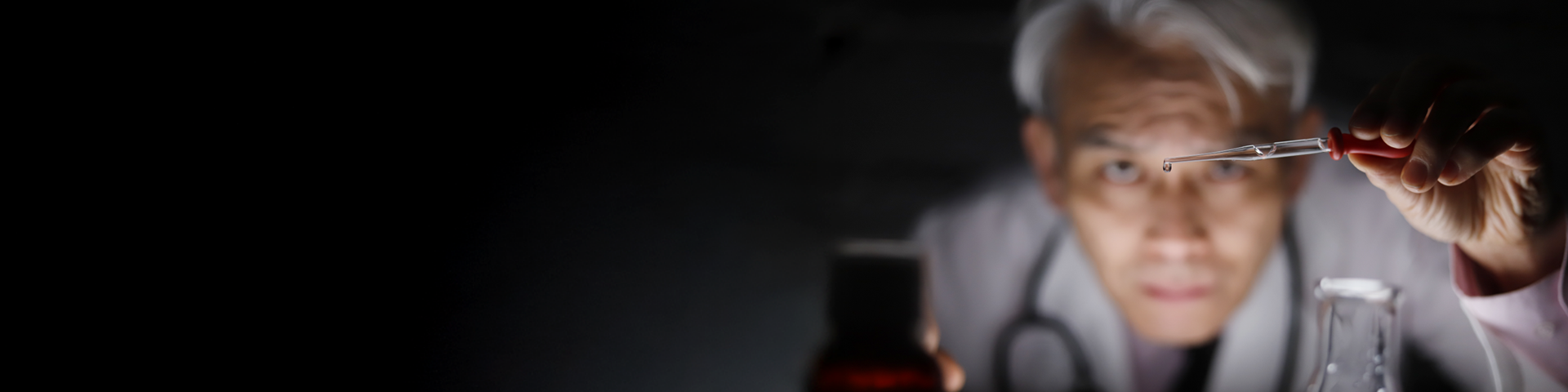 A scientist wearing a lab coat is holding a dropper and dispensing a liquid into a container, with laboratory glassware visible in the background.