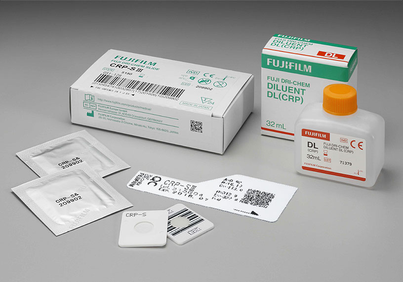 [foto] Caixa de lâminas CRP-SⅢ DRI-CHEM com duas lâminas enroladas em papel alumínio, duas lâminas sem embalagem (vista frontal e traseira) e cartão QC, ao lado da caixa de diluente DL (CRP) e do frasco de diluente DL