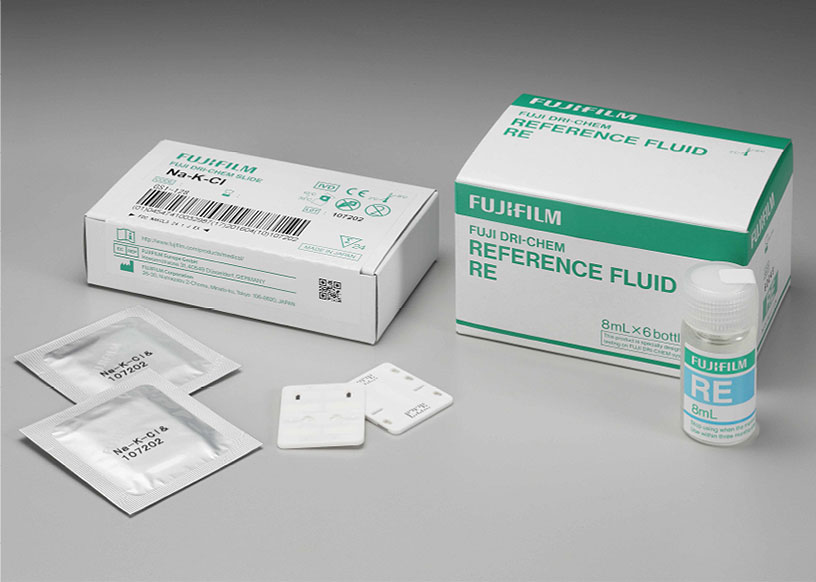 [foto] Caixa de lâminas Na-K-Cl DRI-CHEM com duas lâminas enroladas em papel alumínio, duas lâminas sem embalagem (vista frontal e traseira), ao lado da caixa de fluido de referência RE e frasco de fluido de referência