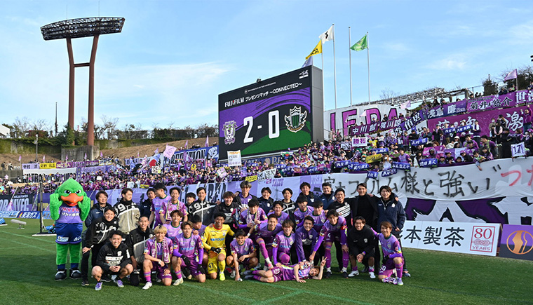 サッカー場の大型ビジョン前で、紫色のユニフォーム姿の参加者らが横一列に並ぶ集合写真。背後のスタンドには多数の観客と応援旗が見える。