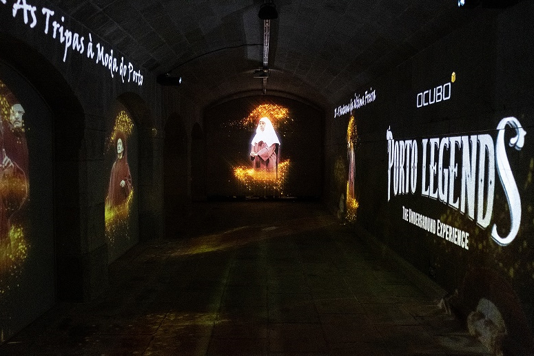 [photo] Woman in red robe and white hood being projected at far end of dark room, with images of actors and words projected unto side walls 