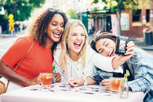 [photo] 3 female friends sitting at a table will sample photo prints while taking a group picture with the Instax Mini LiPlay