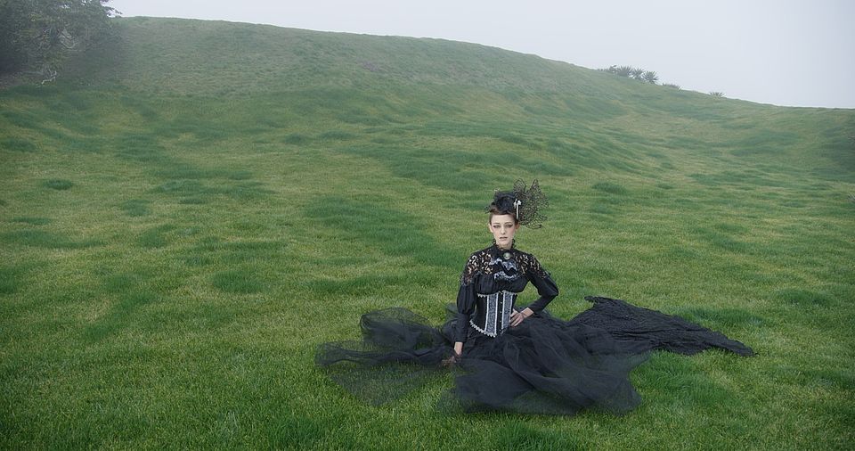 [photo] A wide shot of a lady dressed in a mediaval outfit sitting on a green grass on a cloudy day